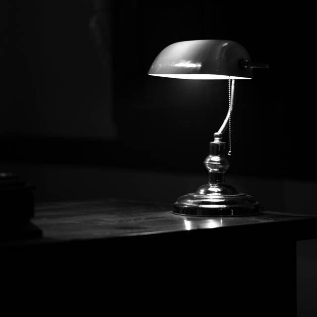 A brass banker’s lamp lit on a dark desk, its light pooling on the polished top while the rest of the room falls away into shadow.