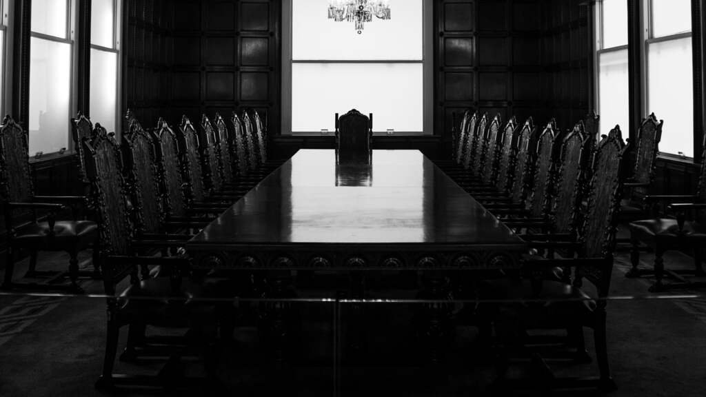 A panelled boardroom after dark: one long polished table, rows of high-backed carved chairs, a chandelier lit above and tall sash windows glowing pale at the far end.