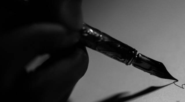 A hand in shadow holding a fountain pen to a blank page, the nib catching the light.