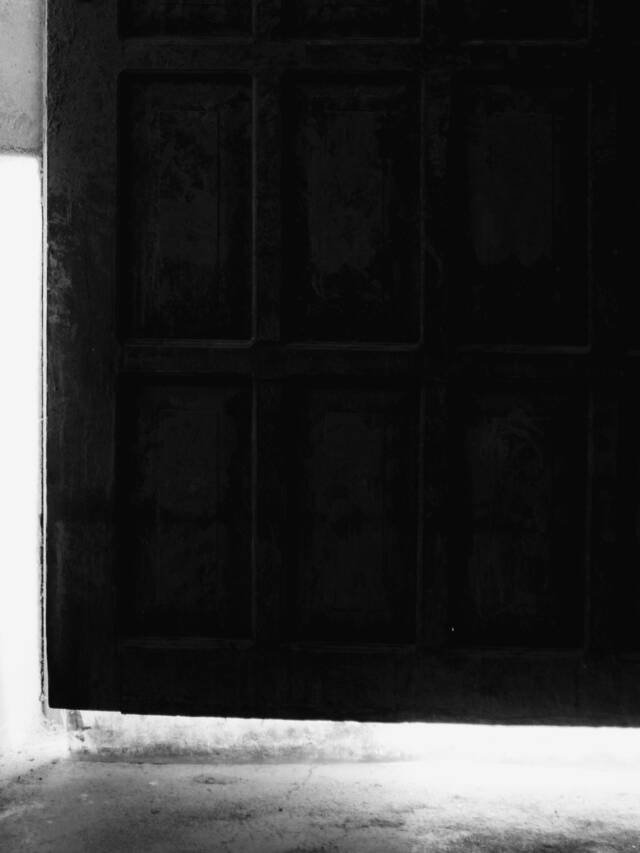 A closed panelled door in a dark corridor, light from the room beyond showing along its edge and under it across the floor.