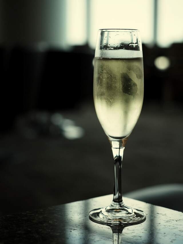 A chilled flute of pale sparkling wine on a dark stone table, window light behind it.