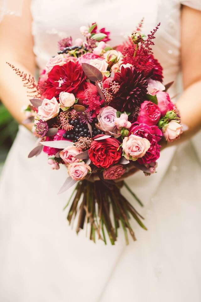 A bridal bouquet of red and blush roses, dahlias and berries, held at the waist against an ivory dress