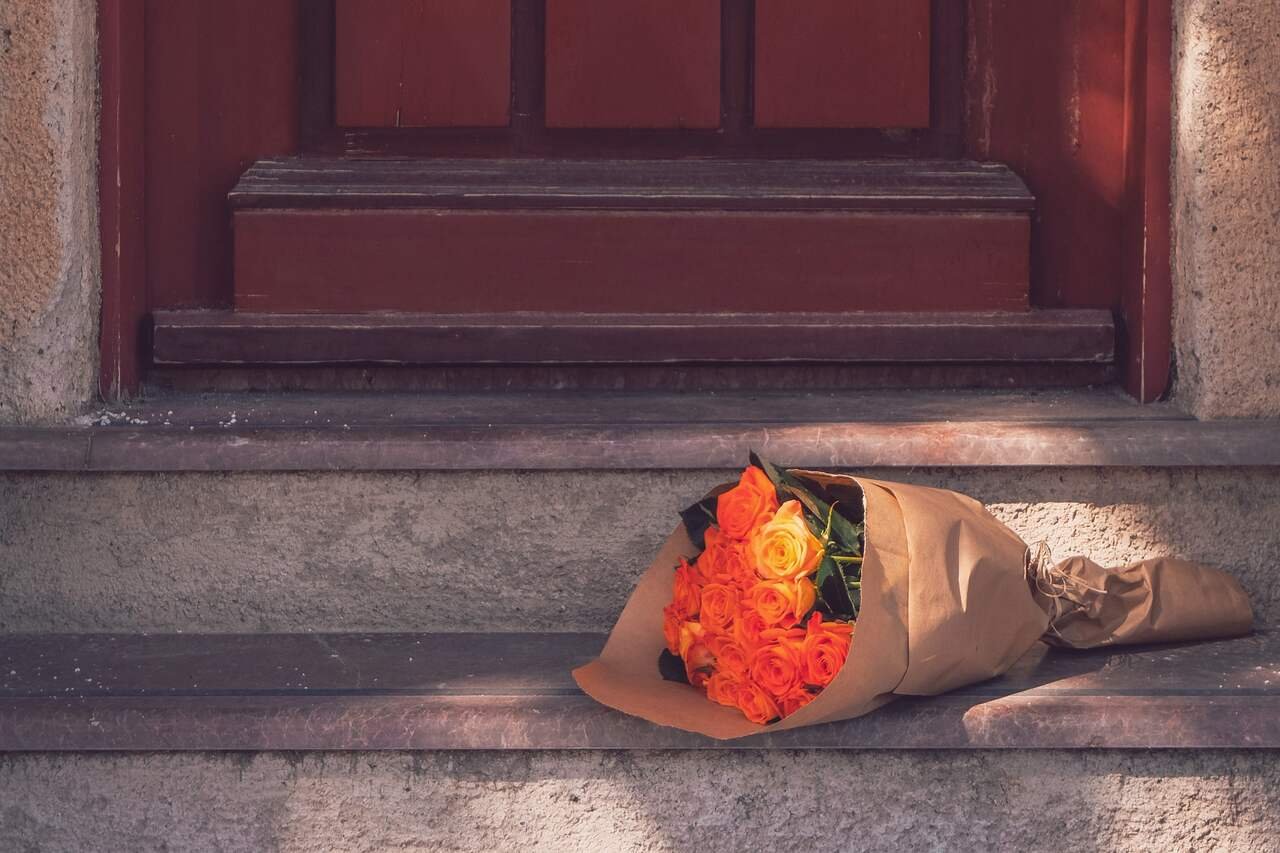 A bouquet of orange roses wrapped in kraft paper, left on a sunlit step at a red front door