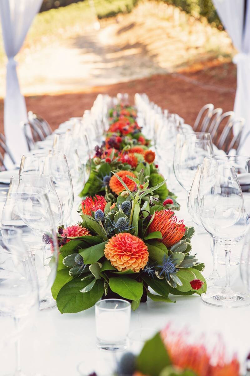 A long reception table set with glasses, and a low run of protea, dahlias and thistle down its centre