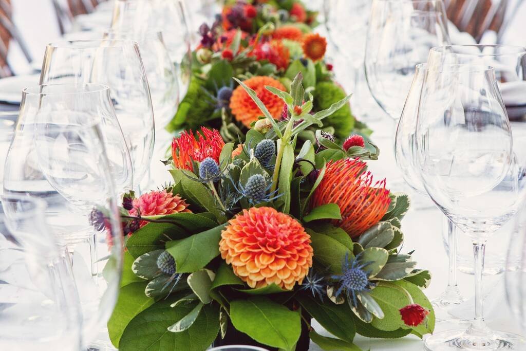 A long reception table set with glasses, and a low run of protea, dahlias and thistle down its centre