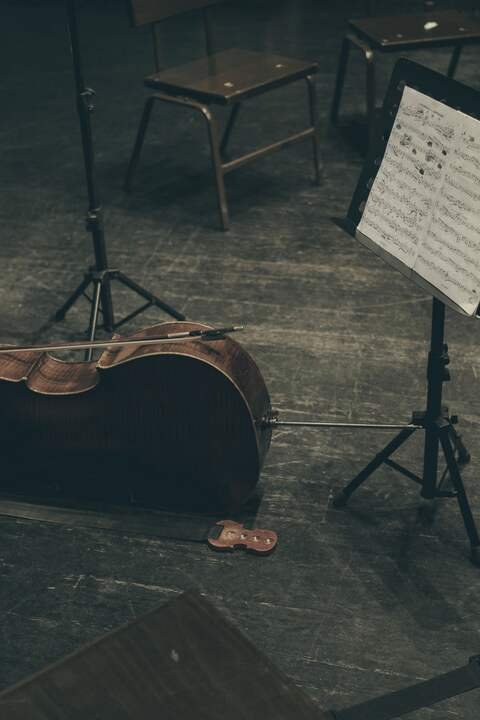 A cello laid on the boards of the practice stage beside a music stand and empty chairs.