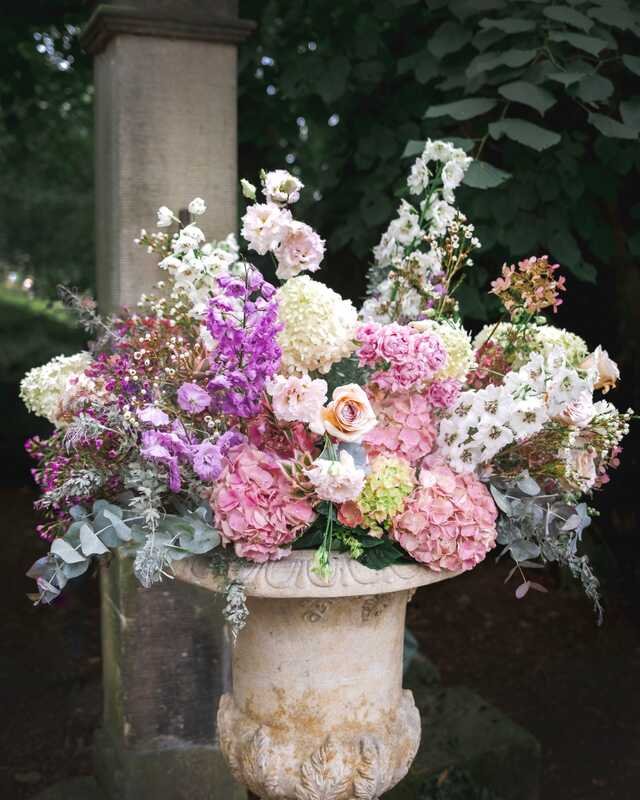 A stone urn spilling with stocks, hydrangeas and roses beside a pillar.
