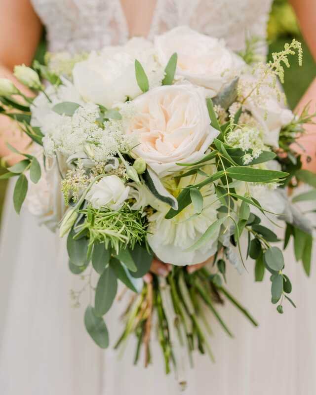 A loose bridal bouquet of cream roses and garden greens, held low against a lace dress.