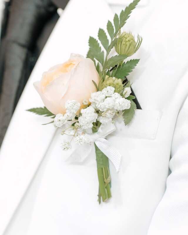 A buttonhole on a cream lapel: one peach garden rose, a sprig of fern, gypsophila and a green scabious pod, tied with white ribbon.
