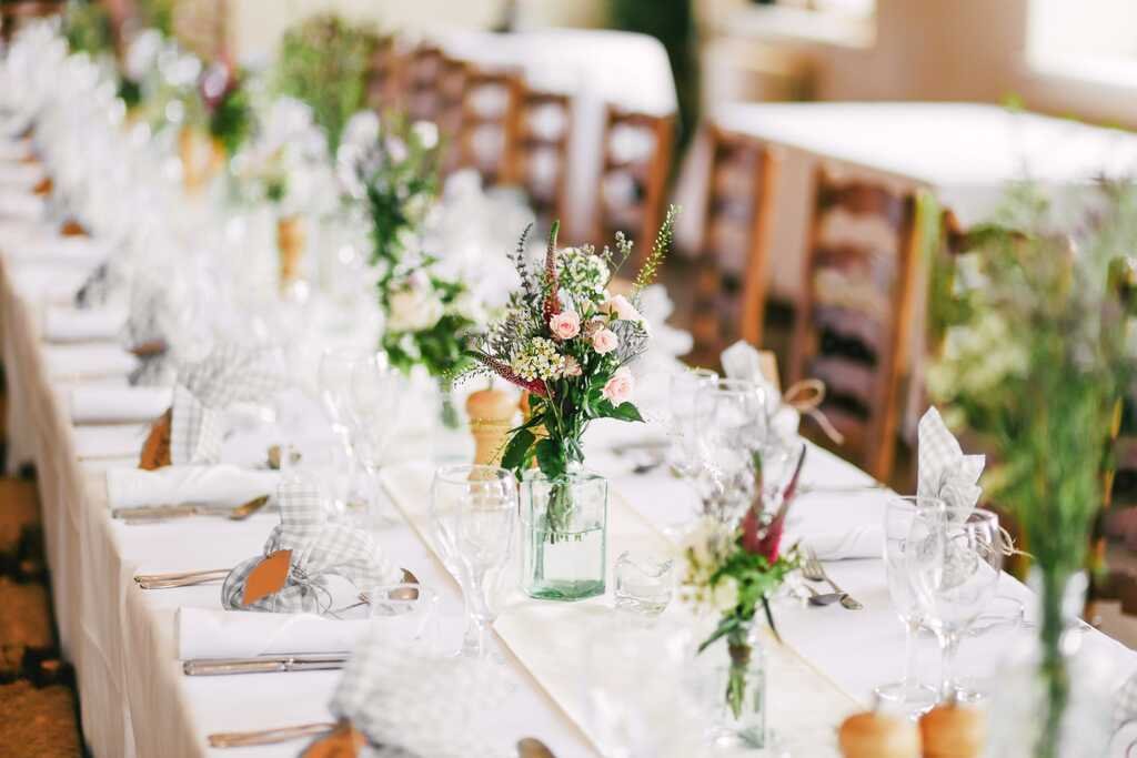 A long wedding table laid in white with oatmeal-check napkins, small glass bottles of garden flowers running its length.
