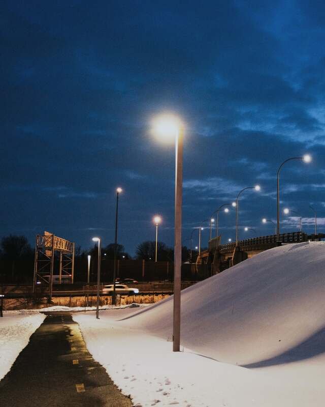 Blue hour after the storm: a cleared path runs wet and black beside a high plowed bank, street lamps still lit.