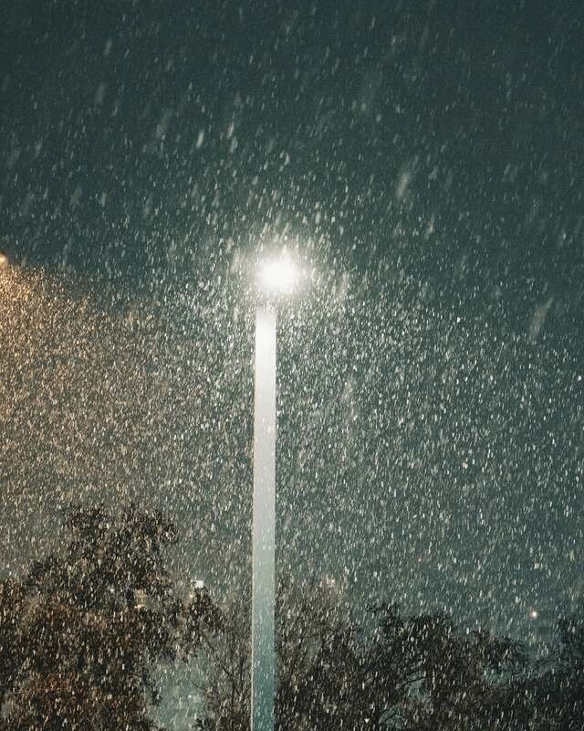 Snow falling thick through a single lamp over a fenced lot at night.