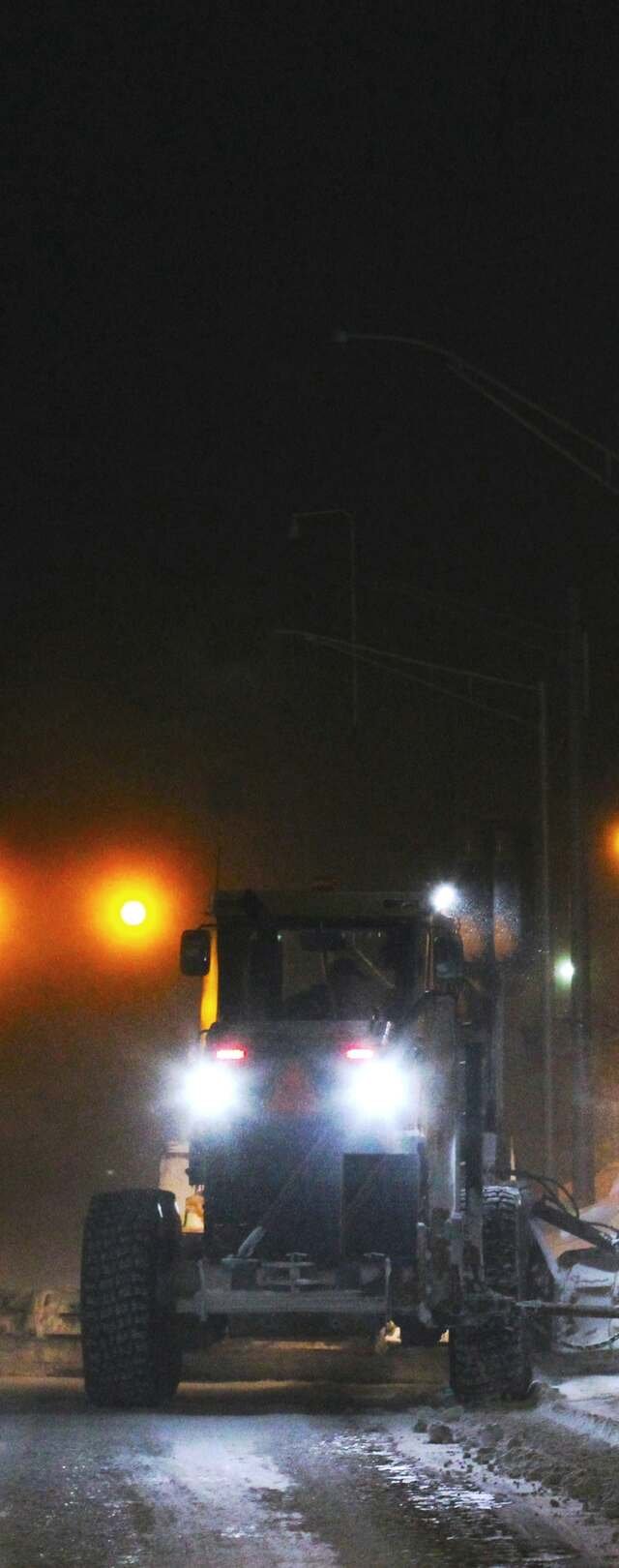 A road grader plows at night under sodium lights, headlights cutting through blowing snow as its blade throws a windrow onto the bank.