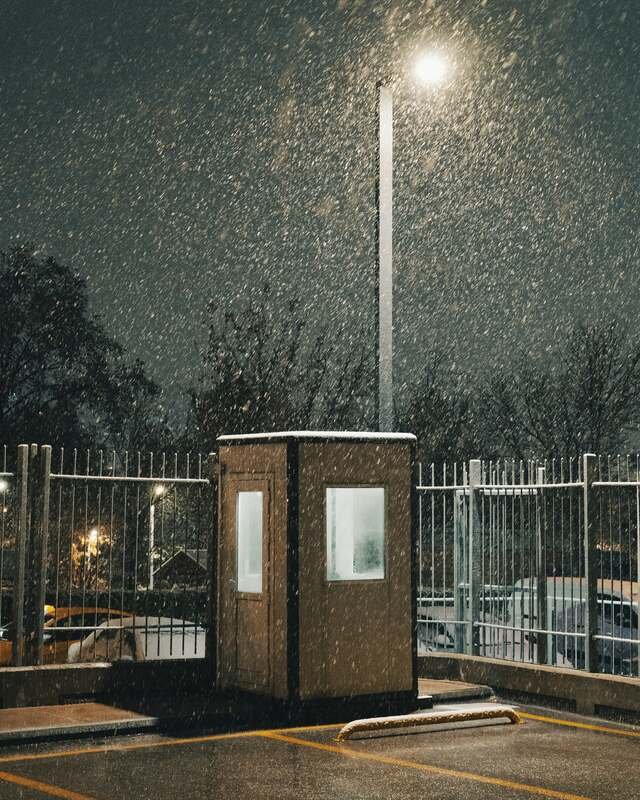 A lot under one lamp at night, the attendant booth and the painted lines going white under fresh snow.