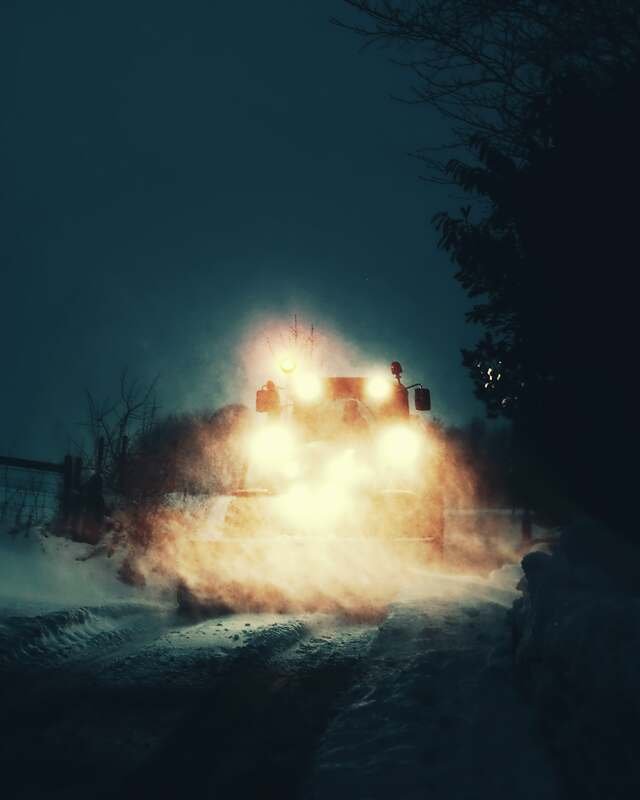 A plow comes head-on through the dark, its lights lighting the spray of snow thrown off the blade.