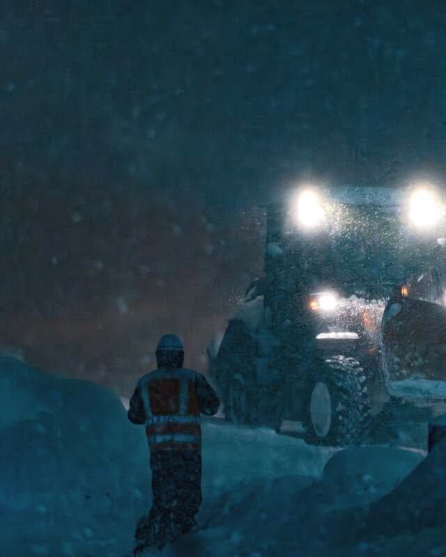 A loader lifts snow onto a pile in a whiteout while a crew member in a reflective vest walks toward it.