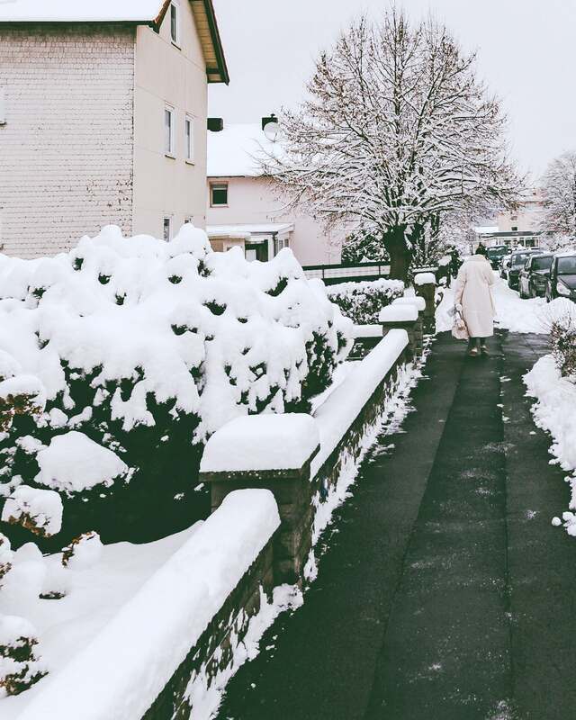 A cleared walkway between snow-covered hedges in a residential street, a neighbour walking away.