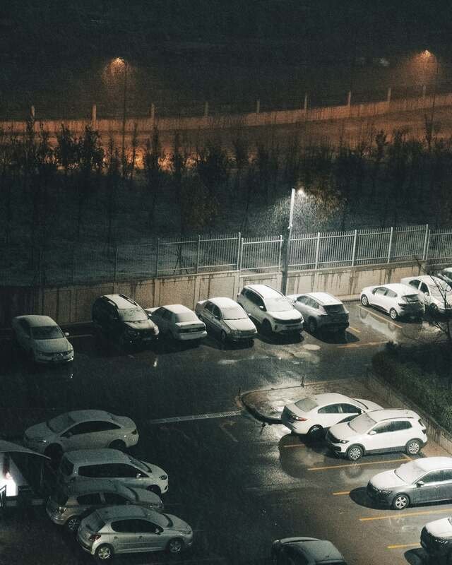 A lot at night in falling snow: parked cars going white while the drive lanes stay wet and black under the lamps.