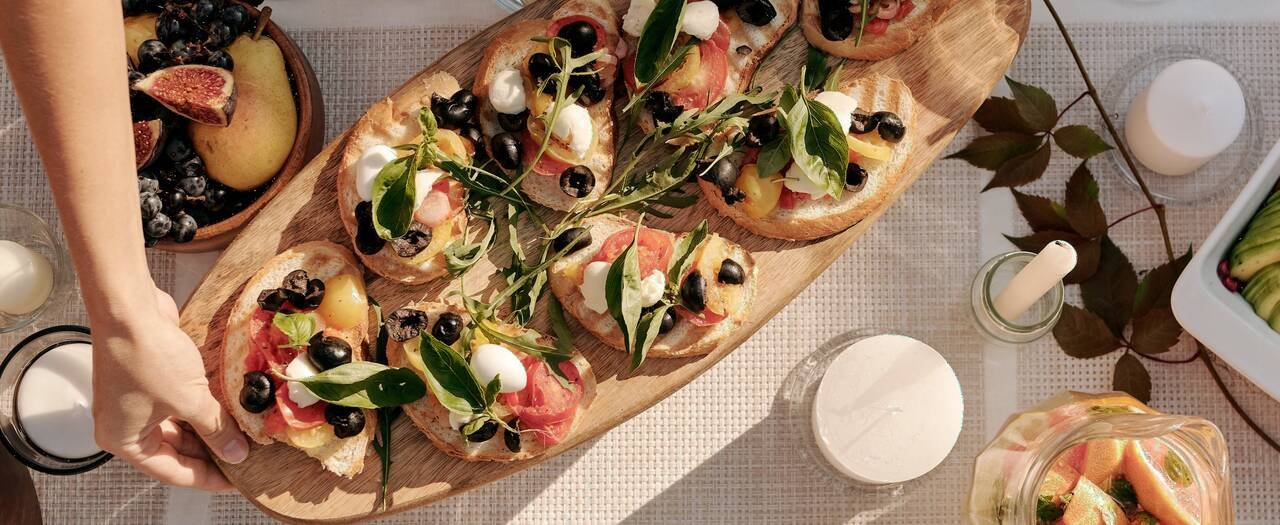 From above, in late sun: a hand sets down a long board of tomato and mozzarella toasts on a linen runner, beside a bowl of figs and a lit candle.