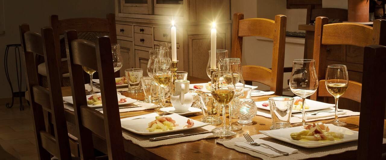 A dining room at night: two candles lit on an old oak table laid for six, white asparagus with ham on every plate, glasses of white wine poured.