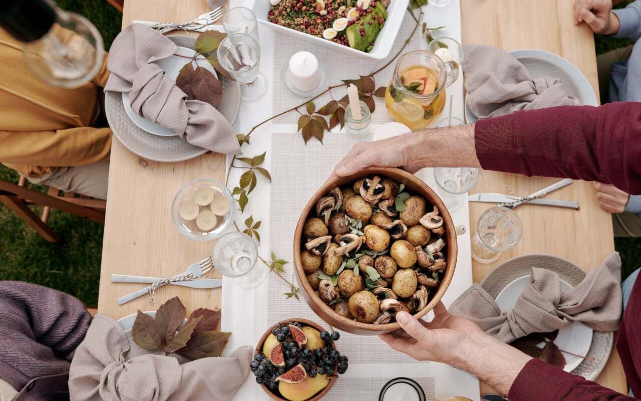 Hands lower a wooden bowl of roasted potatoes and mushrooms onto the middle of the table.