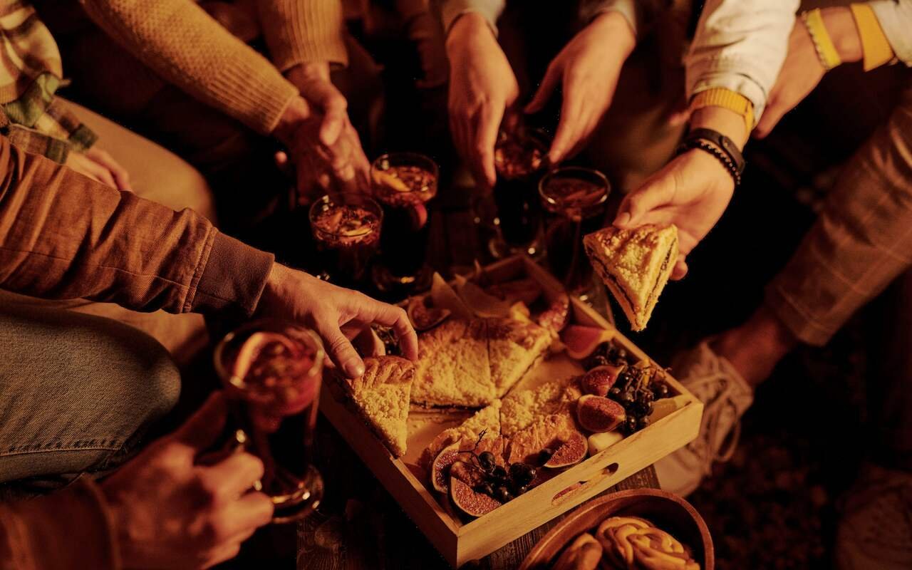 In candlelight, hands reach into a wooden tray of fig and cheese pie cut into wedges, warm drinks among them.