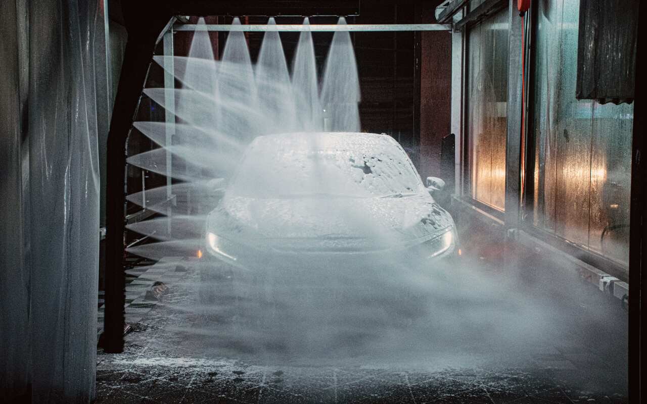 A car under thick white foam in the wash bay, fans of spray falling from the arch above it.
