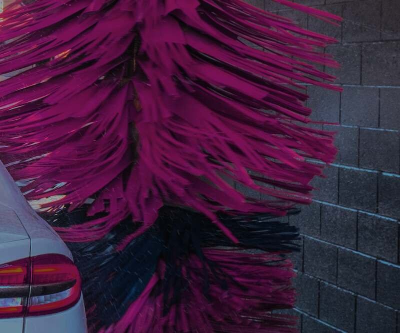 A magenta and blue soft-cloth washer spins down the side of a white car, its tail lamp glowing beside it.