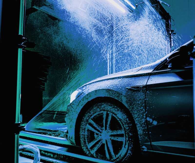 The rinse arch blasts water across the front wing and wheel, spray flaring in the cyan light.
