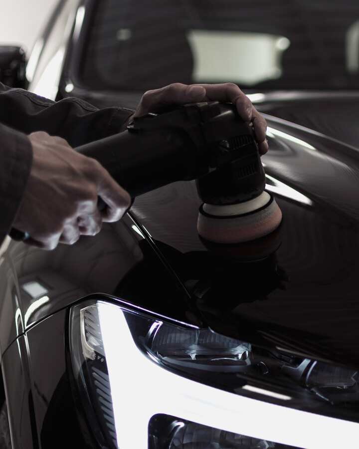 Two hands guide a dual-action polisher across a dark hood, the studio light panels reflected in the paint.