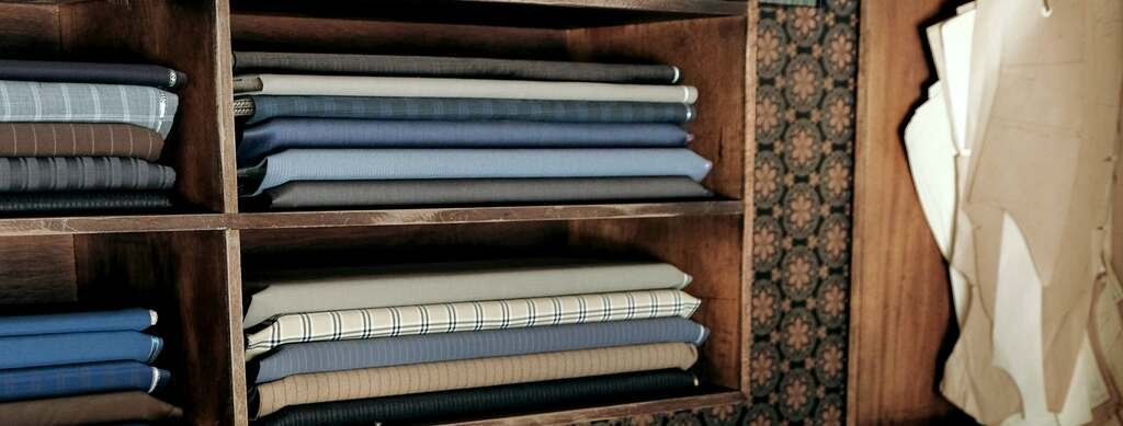 The cutting room: suitings folded on oak shelves, navy, grey, chalk stripe and check, and the paper patterns of past clients hanging beside them.