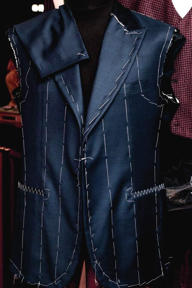 A navy coat at the baste on a dress stand: white tacking down every seam, sleeves not yet set.