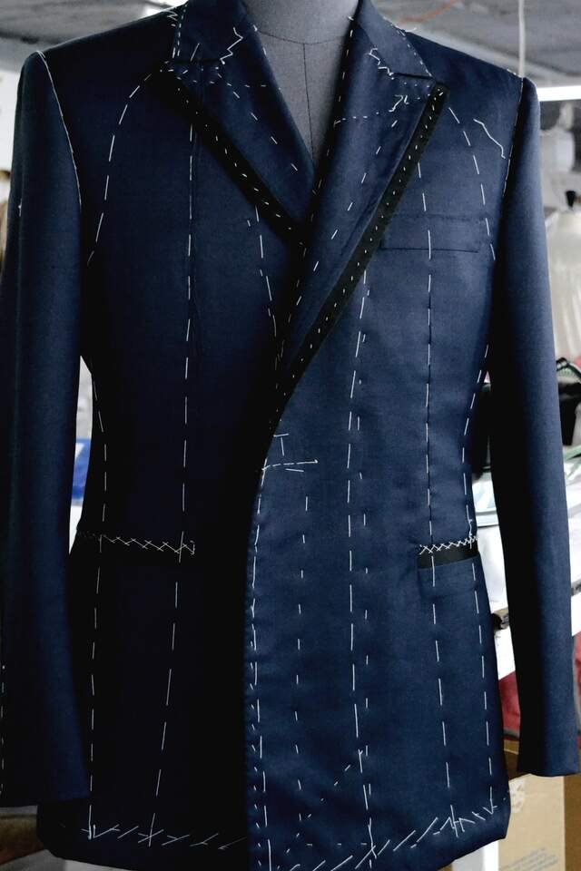 A navy coat at the forward: lapels shaped, pockets in, sleeves set, the last tacking still showing.