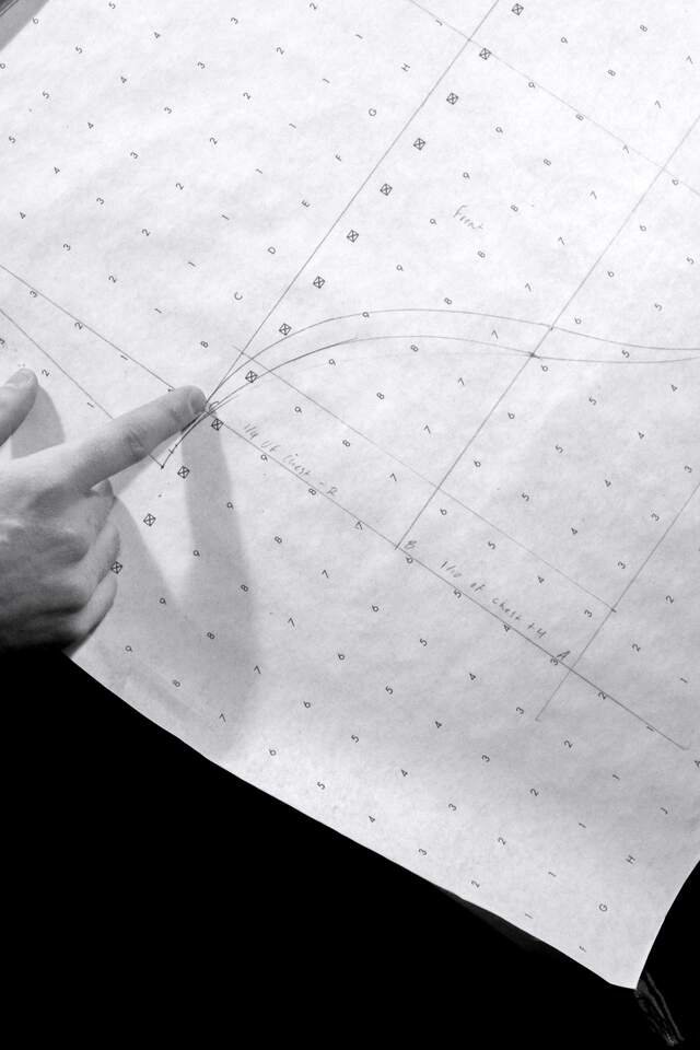 A cutter’s hand on dotted pattern paper, tracing the pencilled armhole curve of a forepart.