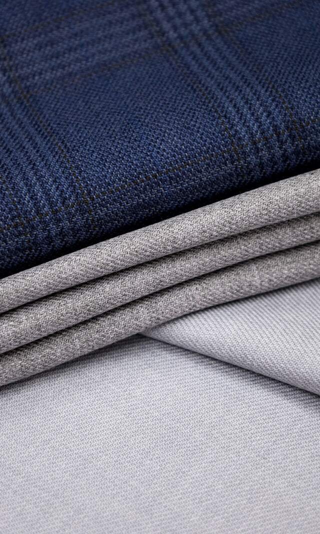 Folded suitings on the cutting table: a navy windowpane worsted over three soft greys, the weave visible in the window light.
