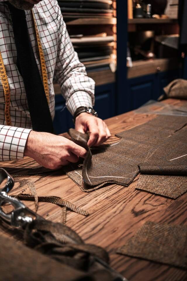 A cutter’s hands on the oak board, lifting a tweed piece marked in chalk, the next pieces already cut beside it.