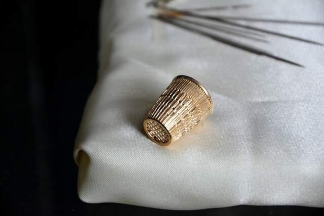 A brass thimble on a fold of pale canvas, a row of long needles above it.