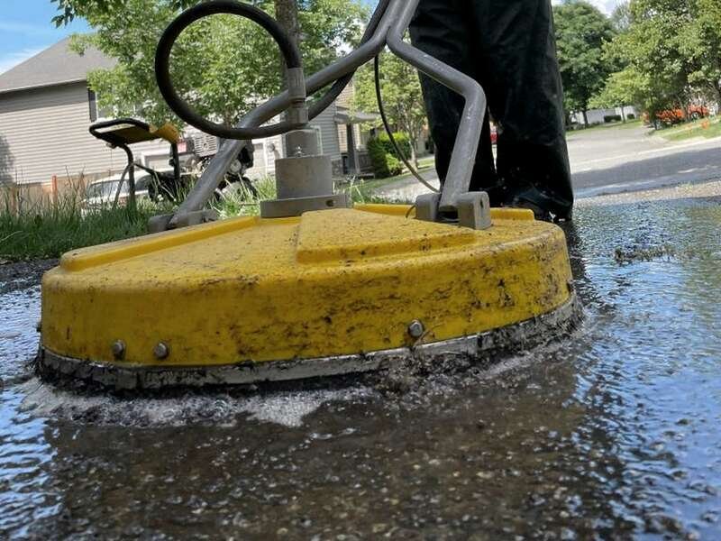 A yellow surface cleaner working across a wet driveway in sunshine, a slurry of lifted grime spraying out from under its rim.