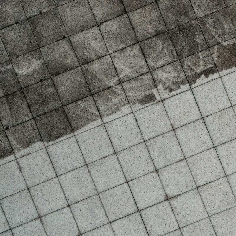 Granite paving tiles seen from above, cleaned half way: dark grime with the swirl of a first pass above, clean pale stone below, divided by one ragged line.
