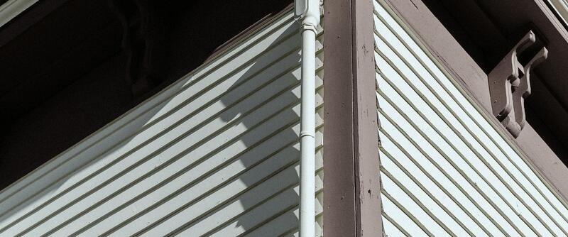 Looking up at the corner of a clapboard house: cream siding in low sun meeting a muted brown corner board and a white downpipe, with carved brackets and deep shadow under the eaves.