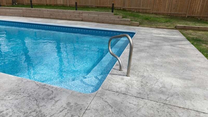 A backyard pool on a bright day: blue water, a steel handrail at the steps and a wide pale concrete deck, with a timber fence behind.