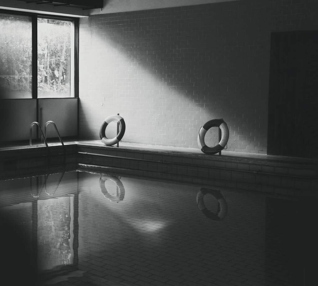 The old plunge hall in the morning: a slant of window light across the tiled wall, and below it water so still it holds the whole room.