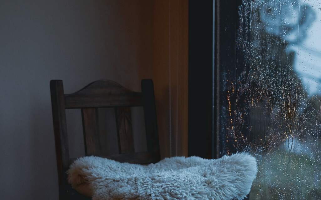 A wooden chair with a sheepskin by the tall window of the quiet hall, rain running down the glass as the evening lamps come on outside.