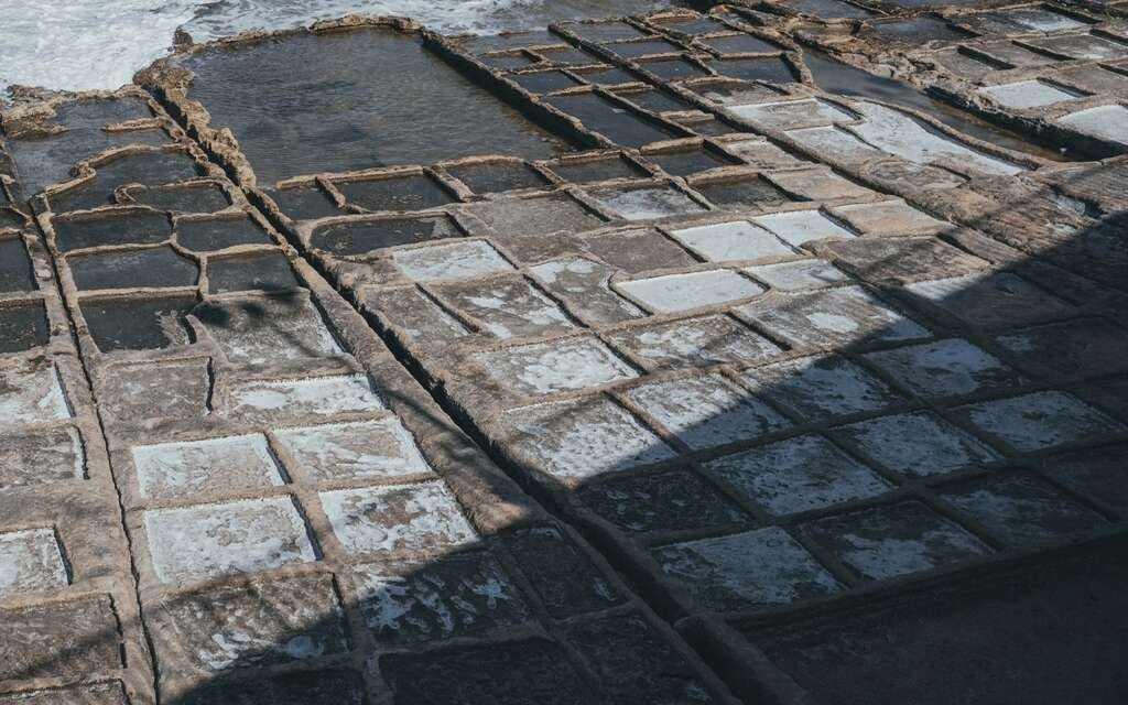 The salt pans below the station: shallow stone basins crusted white with sea salt, the swell breaking just beyond.