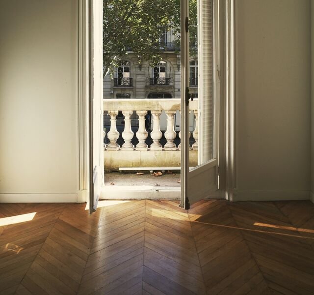 The drawing room of Residence 2.04: tall French windows open onto a stone balustrade, plane trees and limestone houses beyond, afternoon sun lying across a herringbone oak floor.