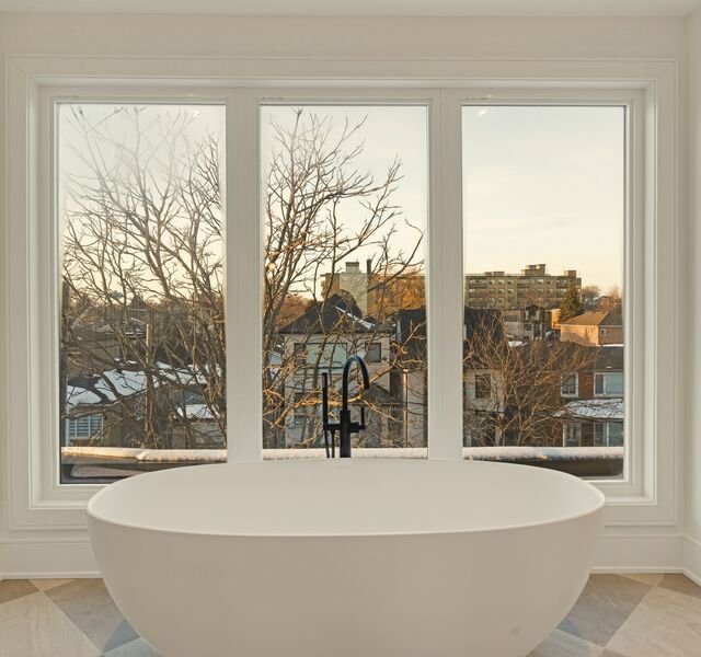 The penthouse bathroom: a freestanding bath in front of a triple window over the rooftops, bare winter trees in low sun.