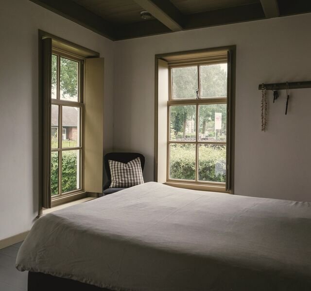 The principal bedroom of Residence 2.04: two deep sash windows with shutters onto garden trees, a made bed in pale linen.