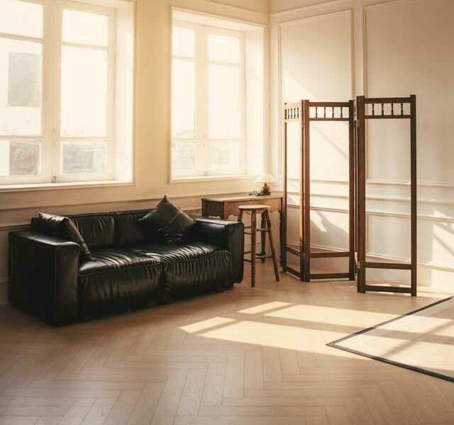 The living room of Residence 1.06: two windows, panelled walls, a dark sofa and low sun across pale oak boards.