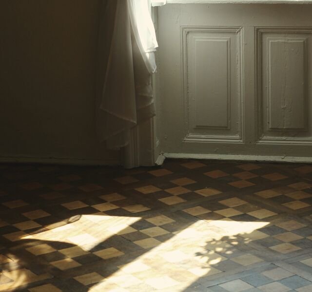 Early light in the principal bedroom of Residence 3.02: a sheer curtain tied back from the window, panelling below, and two squares of sun on the parquet.